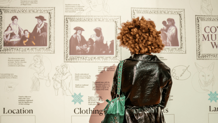 A person with blonde curly hair, wearing a shiny black coat and carry a teal shoulder bag,looking at a display board for the Casta painting exhibition